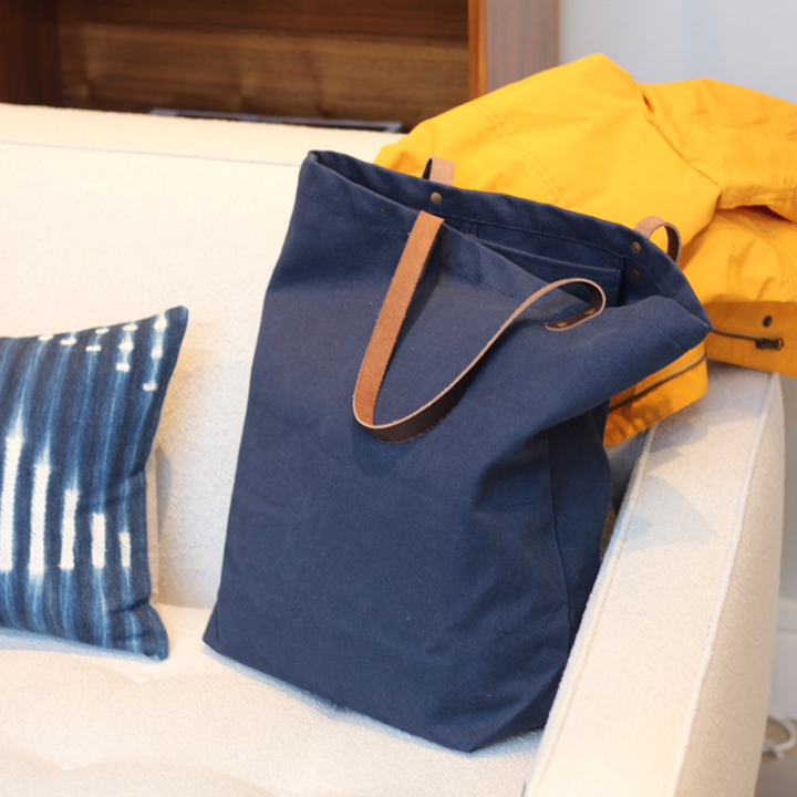 Blue tote bag with brown handles on a white couch with a yellow jacket and blue pillow.