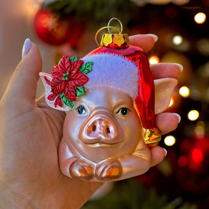 Hand holding a pig-shaped Christmas ornament with a red hat and flower against a blurred festive background.