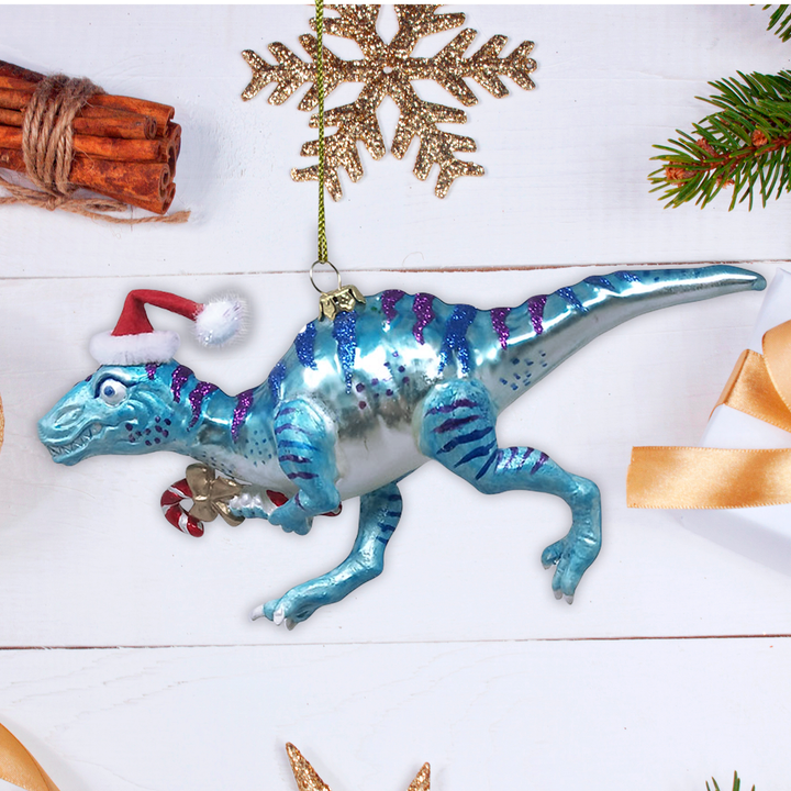 A blue and silver glittery dinosaur Christmas ornament wearing a red Santa hat, displayed on a white wooden background with holiday decorations.