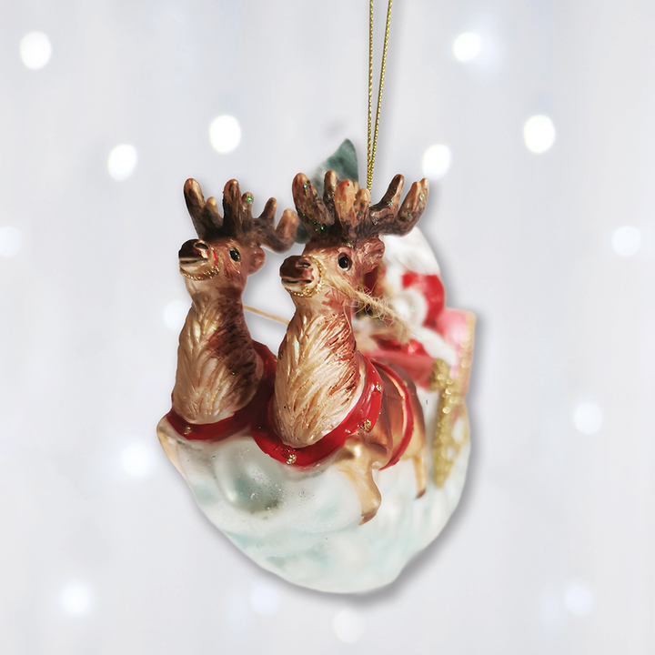 A festive Christmas ornament depicting two reindeer pulling a sleigh on a snowy ground, with a gold hanging string at the top. The reindeer have brown and beige fur with red accents on the sleigh, set against a soft white background with subtle bokeh light effects.