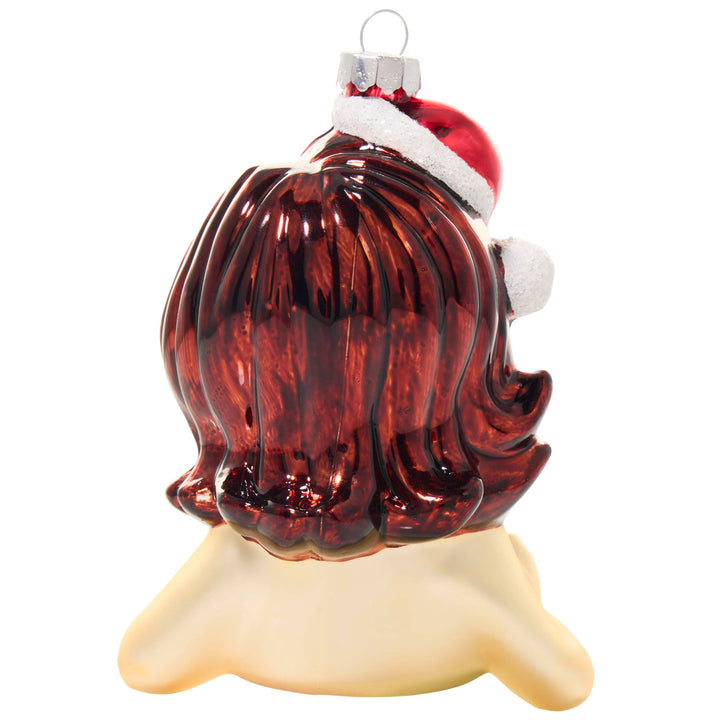 A Christmas ornament shaped like a woman's head with long, glossy dark red hair and a Santa hat, displayed on a cream-colored stand against a white background.