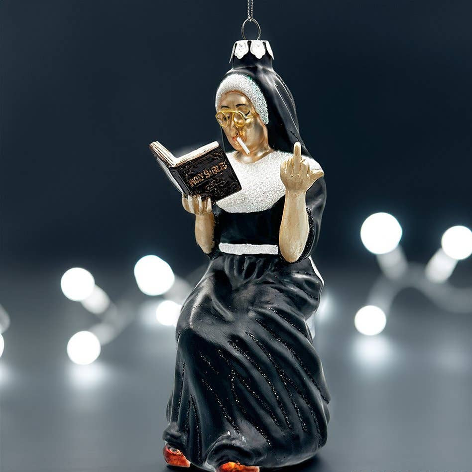 A Christmas ornament shaped like an nun in a black and white habit reading a book, with gold glasses and orange shoes, shown against a dark background with blurred white string lights.