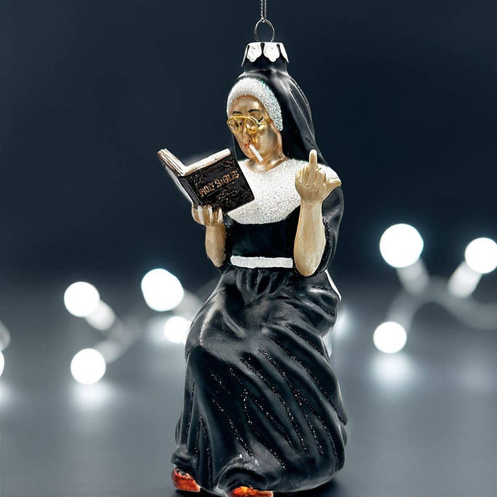 A Christmas ornament shaped like an nun in a black and white habit reading a book, with gold glasses and orange shoes, shown against a dark background with blurred white string lights.