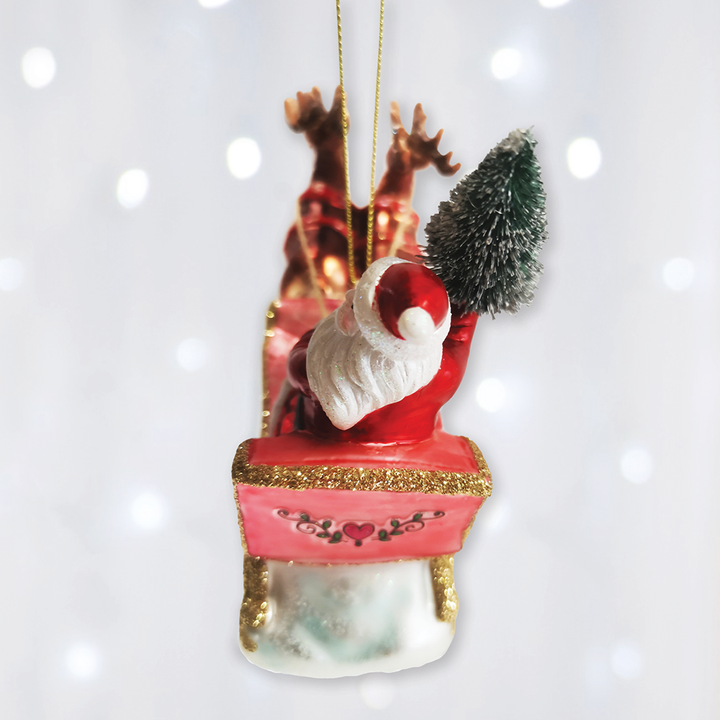 A festive Christmas ornament depicting Santa Claus sitting in a red sleigh with gold glitter trim, accompanied by two brown reindeer and a small green tree, hanging against a soft white background with bokeh light effects.
