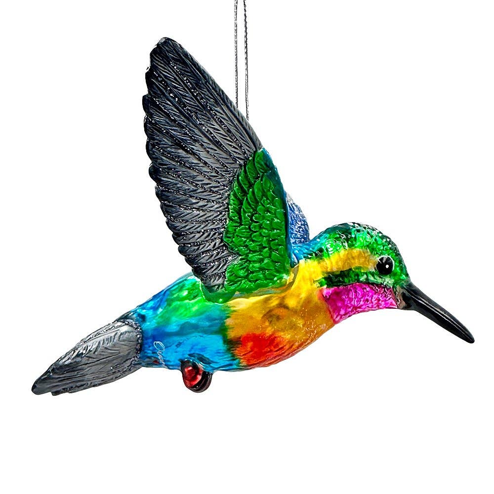 A vibrant, multicolored hummingbird ornament shown in profile against a solid white background. The ornament features a glossy, rainbow-like finish with green, blue, yellow, orange, and pink tones, black wings, and a small red accent near the tail.