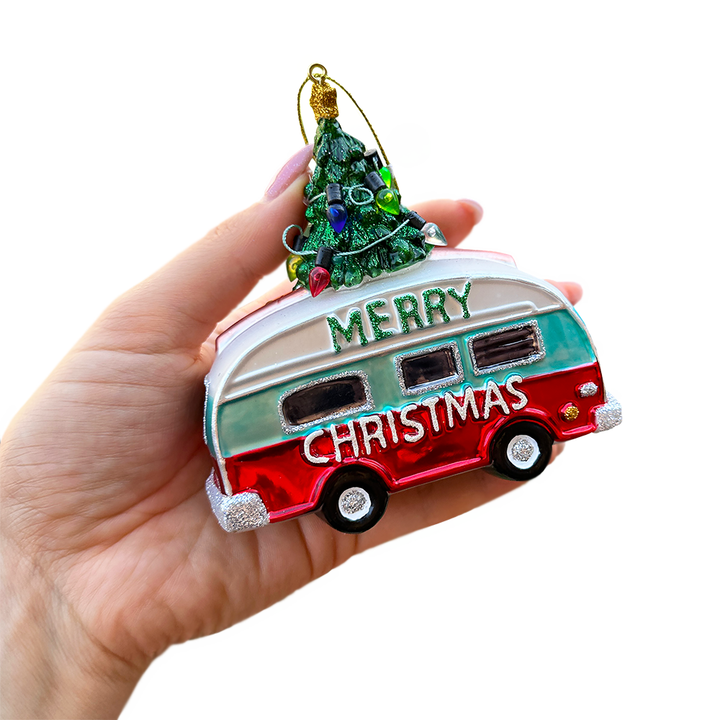 A hand holding a Christmas ornament shaped like a retro camper van with a small decorated tree on top. The ornament has 'MERRY CHRISTMAS' written on it in white lettering, with red, teal, and white colors, and silver glitter trim, set against a plain white background.
