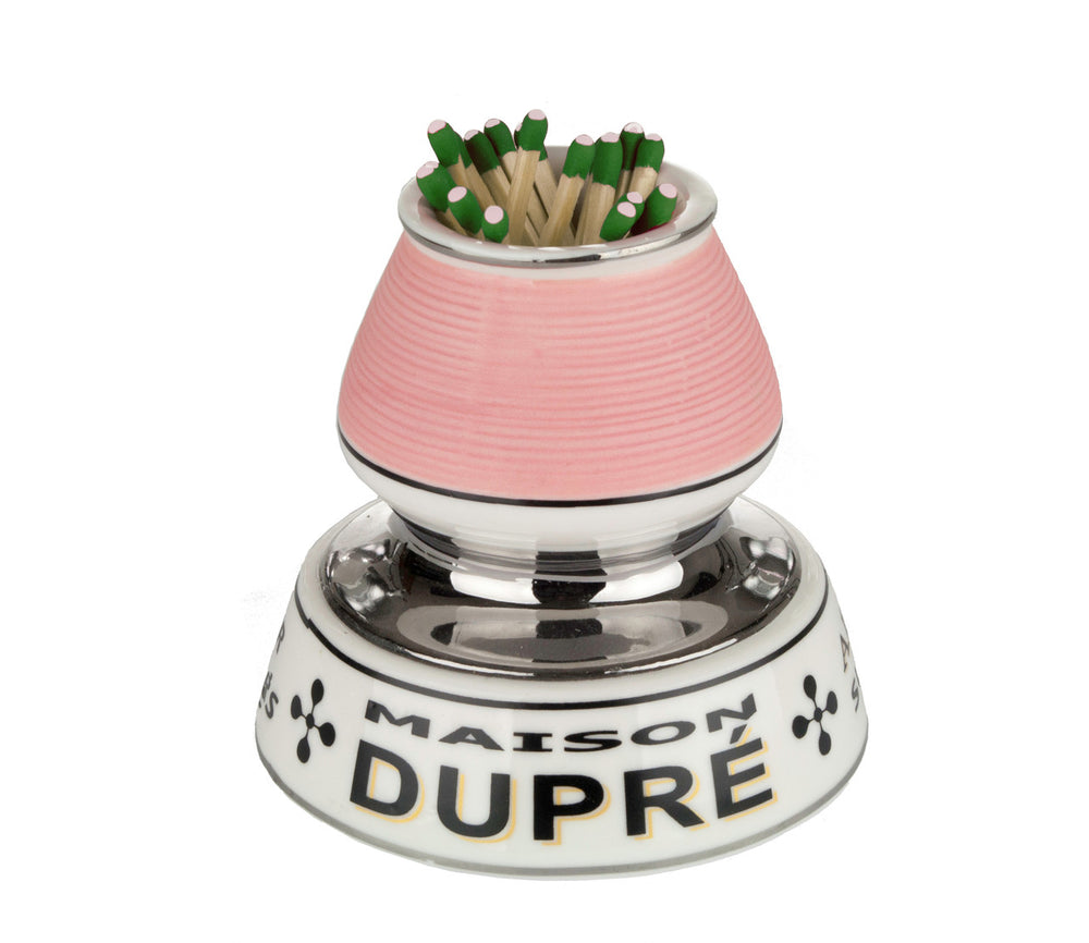 A small cylindrical ceramic match striker, painted white with subtle hand‑painted lines and chrome accents. It stands about 3.5 inches tall with a hollow cavity at the top holding red‑tip “strike anywhere” matches, while its ridged side or base provides a surface for striking matches. The striker is shown against a plain white background, highlighting its compact shape and smooth glossy finish — a vintage‑style piece evoking a 19th/20th‑century European bistro aesthetic.