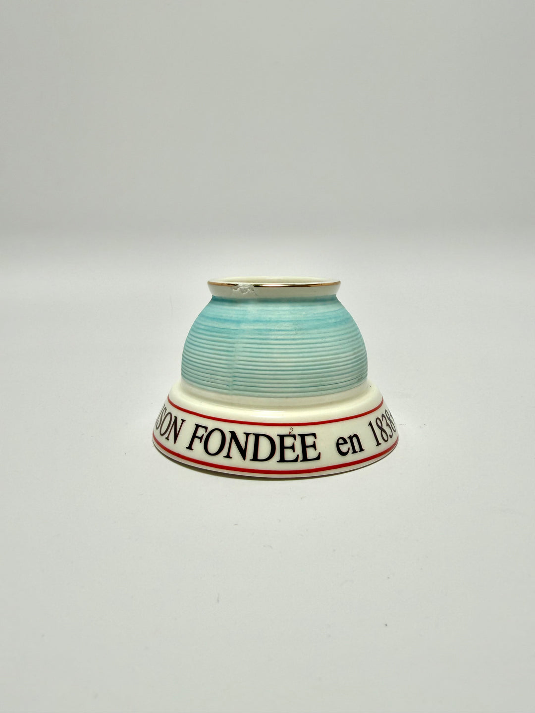 Close-up of a ceramic match striker with a light blue exterior featuring horizontal ridges, a white interior, and a thin gold rim and a small chip.