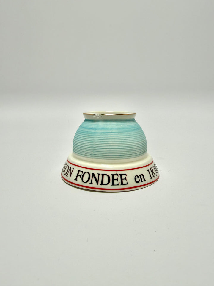 Close-up of a ceramic match striker with a light blue exterior featuring horizontal ridges, a white interior, and a thin gold rim and a small chip.