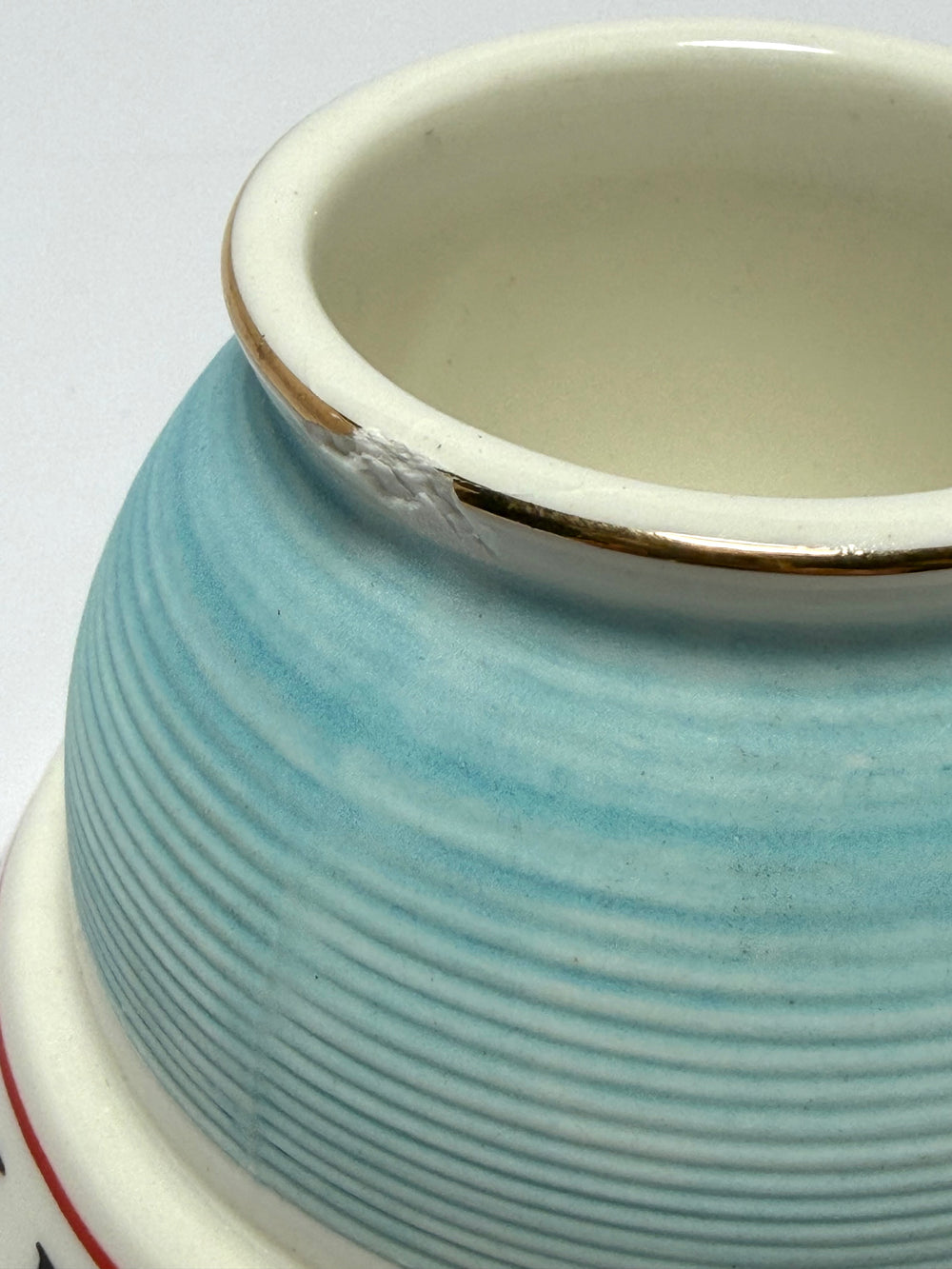 Close-up of a ceramic match striker with a light blue exterior featuring horizontal ridges, a white interior, and a thin gold rim and a small chip.