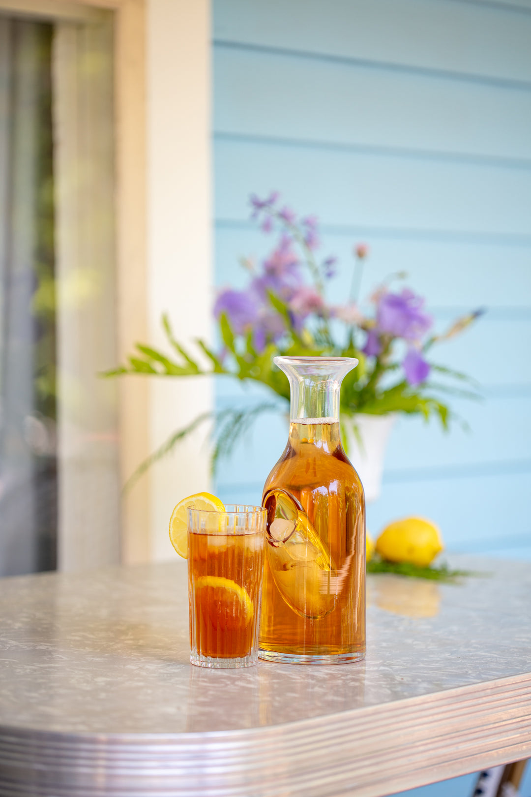 Ice carafe made of glass with a reservoir for ice, shown with a clear glass with iced tea and lemon slices, on a retro Formica and chrome table on a covered porch with light blue clapboards behind the table with a flower arrangement of irises.