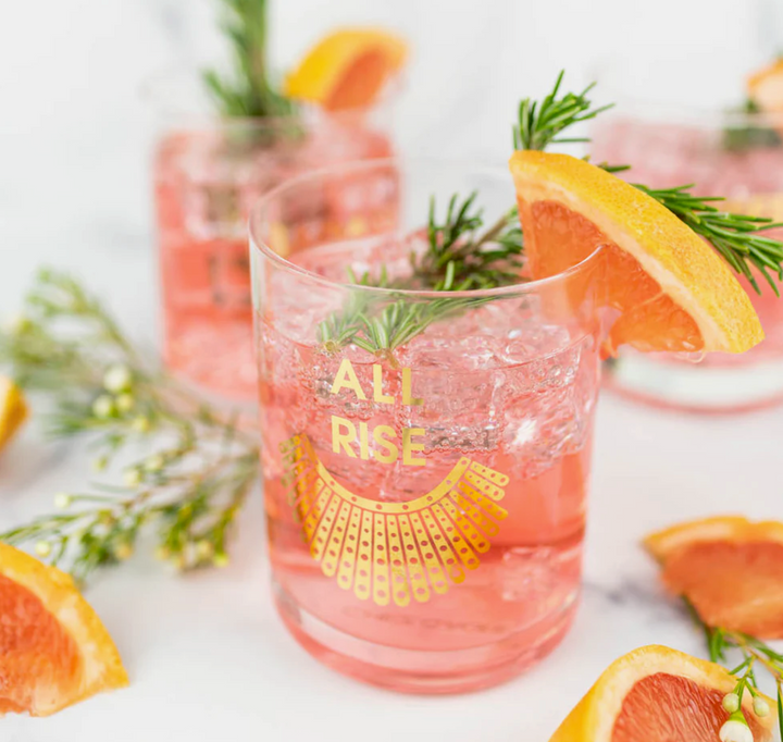 Pink cocktail in a rocks glass with 'All Rise' in gold foil print, garnished with orange slices and rosemary, on a white background.