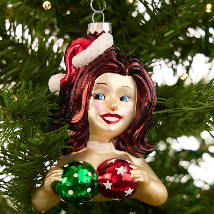 A vintage-style Christmas glass ornament shaped like a woman with red hair, a Santa hat, and two shiny decorative balls, hanging on a green Christmas tree.