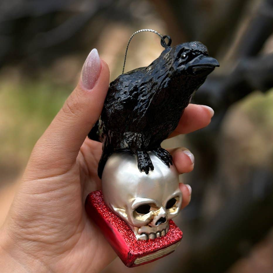 Decorative ornament with a black bird on a skull on a red book base held by a hand.