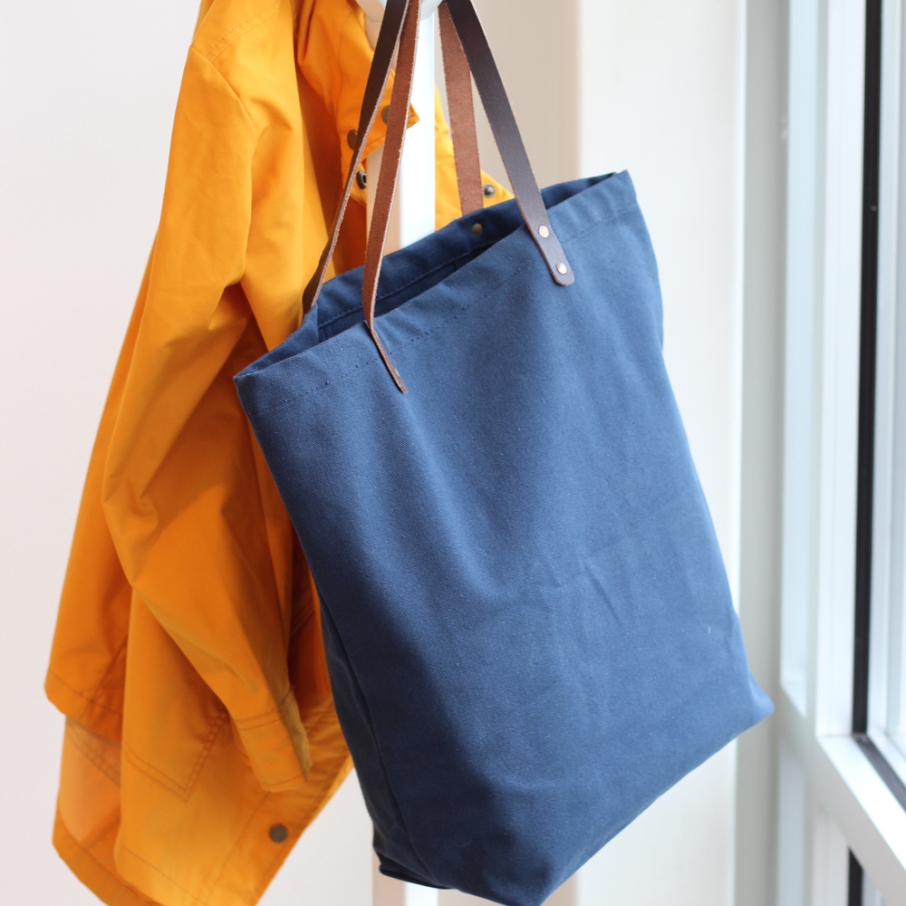 Blue tote bag with brown handles hanging on a yellow jacket against a white wall.