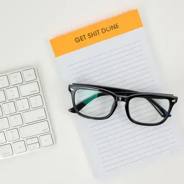 Eyeglasses on a notebook with 'GET SHIT DONE' cover, next to a keyboard on a light background