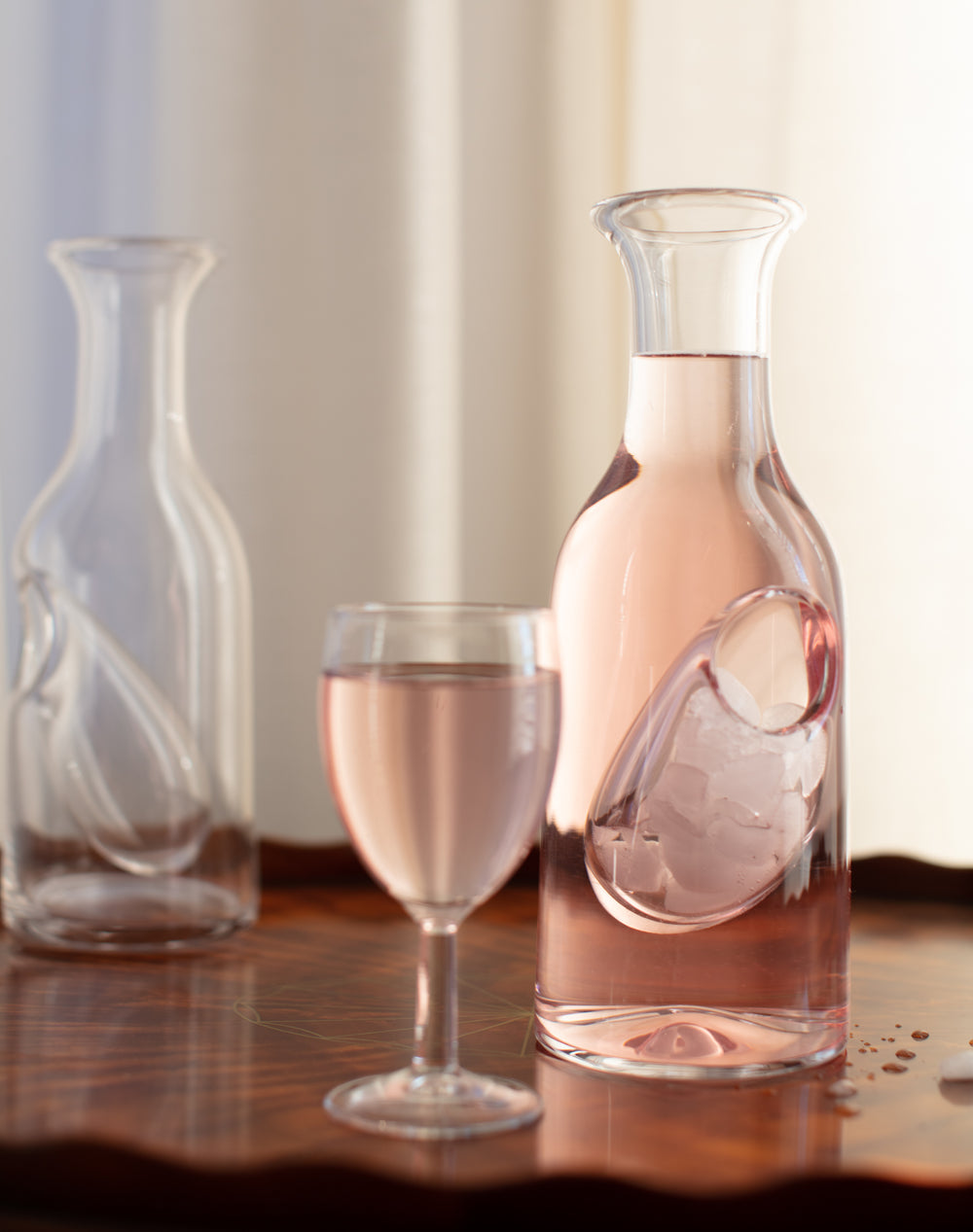 A clear glass ice reservoir carafe with a glass cup next to it, filled with a pink liquid, likely to be ice or a flavored drink, on a wooden surface.