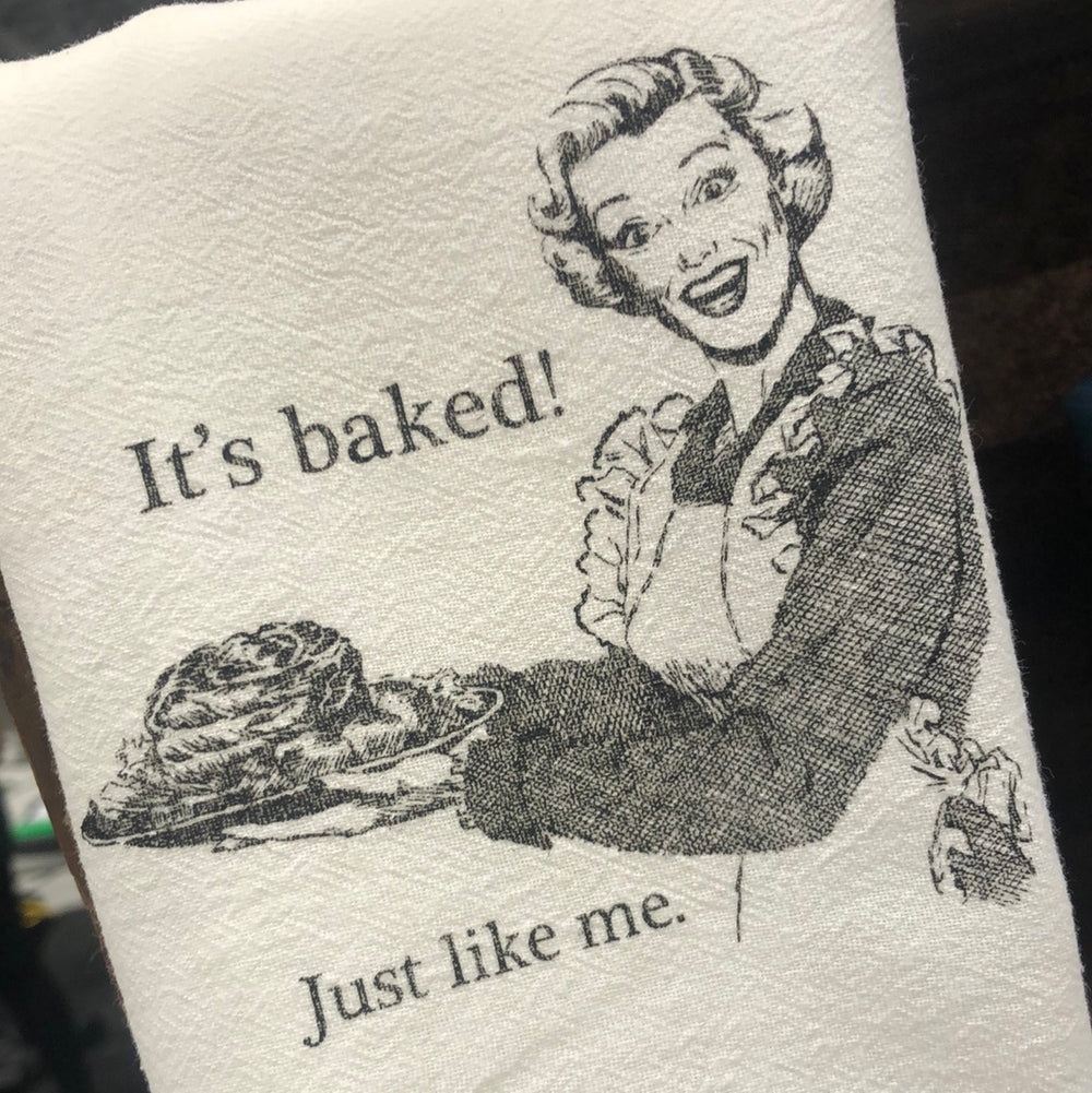A white cotton tea towel with a black and white illustration of a woman and the text 'It's baked! Just like me.'